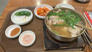 【画像】釜山駅で食べたクッパ。やや辛めのスープに、ご飯と豚（と思われる）腸詰め、野菜が煮込まれている。付け合わせとして、生のピーマンとニンニクが添えられていた。どうやらそのまま食べるもののようである。非常に美味しく、何よりも体が自然に求めていると感じた。